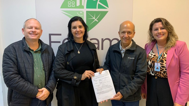 Quatro pessoas em pé diante de um painel com a logomarca da Fepam. As duas pessoas no centro da imagem seguram o termo que foi assinado. Da esquerda para direita: Da esquerda para direita: Henrique Lagemann, assessor da Sedur que representou o secretário Rafael Mallmann; Caroline Techio, bióloga da Metroplan; Renato Chagas, diretor-presidente da Fepam, e Gabriela Goldani, diretoria da Metroplan.