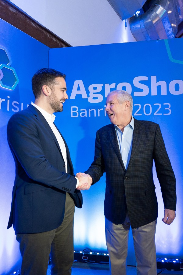 Leite aperta a mão de Coutinho, com um fundo azul onde está escrito Agroshow. Ambos sorriem.