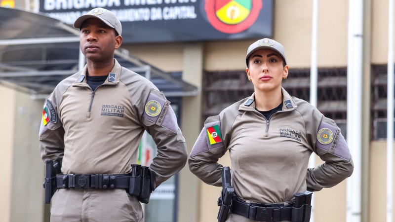 Em ambiente aberto, um brigadiano e uma brigadiana estão postados de pé. Os dois trajam boné da mesma cor do uniforme, que é bege.