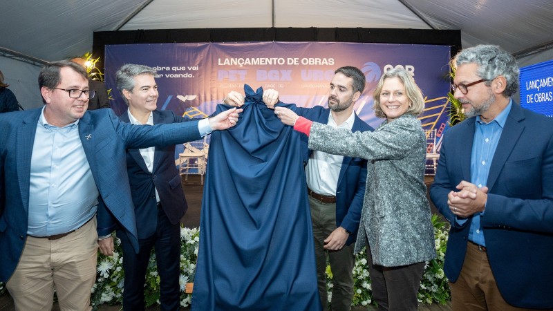 Ao centro e diante de um painel informativo sobre o lançamento das obras nos três aeroportos, Eduardo Leite e a prefeita de Pelotas retiram o pano que está sobre a placa que marca o início das obras. Ainda há mais três pessoas participando do ato que aparece na imagem.