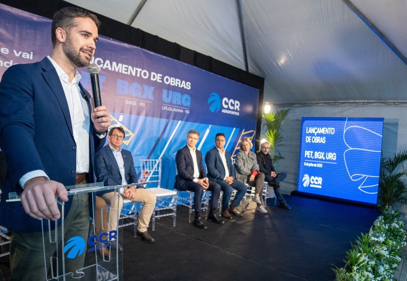 Eduardo Leite à esquerda da imagem discursando ao microfone. Na imagem, há mais cinco pessoas sentadas à direita de leite compondo a mesa. Elas estão à frente do painel de lançamento das obras e atentas à fala do governador.