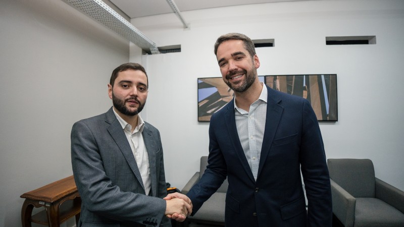 O governador Eduardo Leite, de casa azul marinho e camisa branca, e o vice-governador Gabriel Souza, de casaco cinza e camisa branca, apertam as mão. Ao fundo, poltronas cinzas e um quadro com uma fotografia na parede. 
