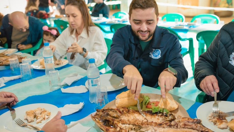 Gabriel souza, vestindo uma jaqueta azul com o símbolo da Feira Nacional do Peixe, sorri enquanto corta com garfo e faca um pedaço de uma tainha assada que repousa sobre um refratário em uma mesa com toalhda azul em listras largas nas cores azul claro e escuro. Ao lado de Gabriel, sua esposa, Talise. Também sobre a mesa, garrafinhas de água mineral.