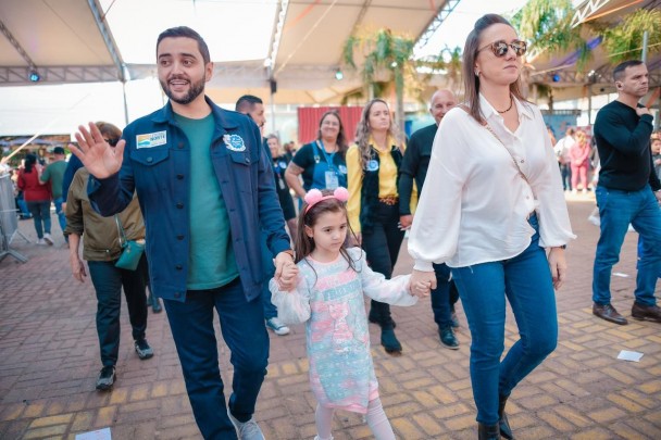 Gabriel Souza, vestindo uma jaqueta azul, camiseta verde e calça jeans, acena com a mão esquerda, enquanto caminha nos espaços da Feira do Peixe. Com a mão direita, segura a mão da sua filha, Dora, que veste um vestido branco e uma tiara rosa com dois pompoms. Com a outra mão, Dora segura a mão da mãe, Talise, que caminha ao lado vestindo blusa branca e calça jeans. Atrás dos três, outras pessoas que visitam a feira em Tramandaí.