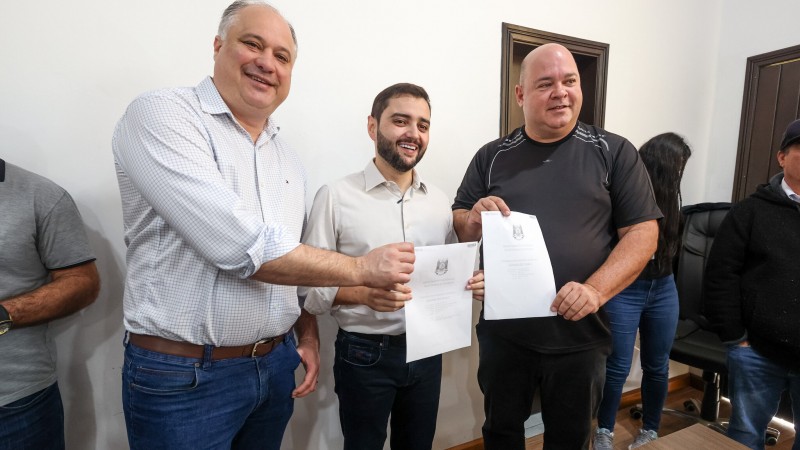 Numa sala fechada, Luciano Silveira, Gabriel Souza e Ique Vedovato estão em pé, da esquerda para a direita. Ele exibem duas folhas com assinaturas e sorriem.