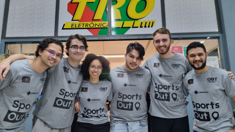 Cinco jovens, trajando a camisa cinza da instituição, posam para a foto. Eles estão em um ambiente fechado, com os braços ao redor dos ombros uns dos outros, e sorriem para a câmera