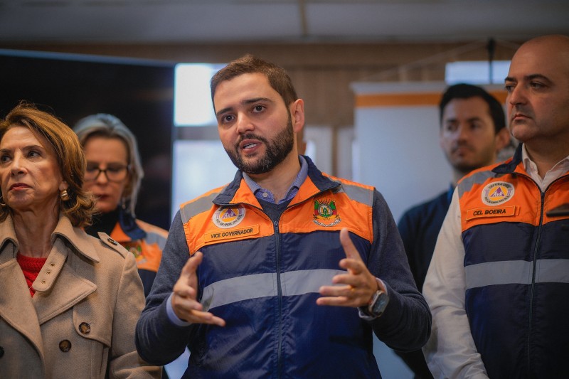 Gabriel Souza, trajando o colete da Defesa Civil, está em pé, falando e gesticulando. Ao seu lado e atrás, outros representantes do governo aparecem com expressão séria.