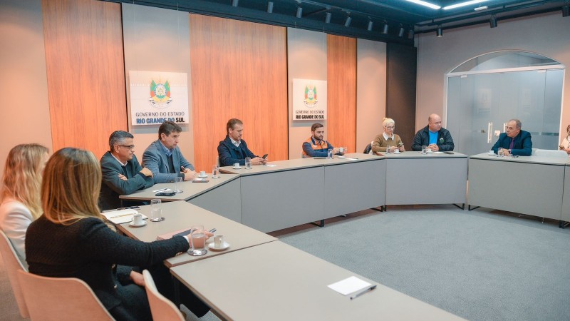 Na sala fechada, várias pessoas estão sentadas às mesas dispostas de forma rente às paredes. Nas paredes, aparece a logo do governo do RS