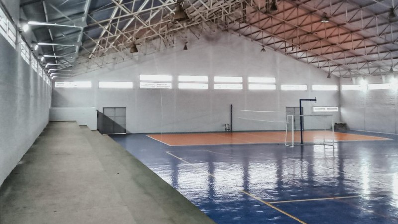 Ginásio esportivo, com quadra de cores azul e laranja e arquibancadas cinzas. Paredes são brancas.