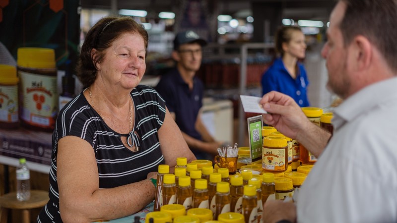 Num ambiente de feira, uma senhora sorri em pé atrás de um balcão com vários vidros de mel. O secretário Santini estão no canto direito, segurando uma cartão de visitas na mão.