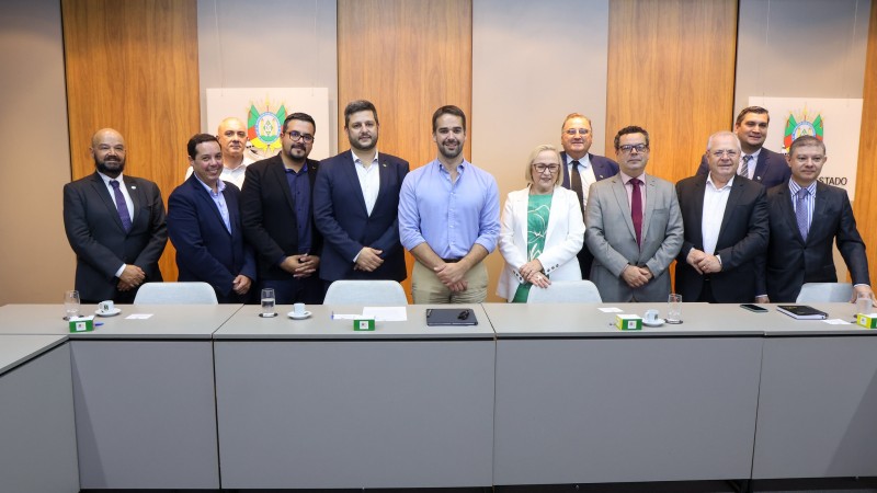 Numa sala de reuniões, Eduardo Leite está de pé, ao centro, tendo a seu lado a secretária da saúde, Arita, e do turismo, Vilson Covatti. Ao lado deles, há vários outros homens também de pé, trajando terno. Todos posam para a foto.