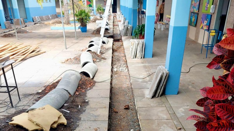 No pátio de uma escola, estão dispostos diversos materiais de obras. Aparecem pilastras azuis e quadros coloridos nas paredes ao fundo.
