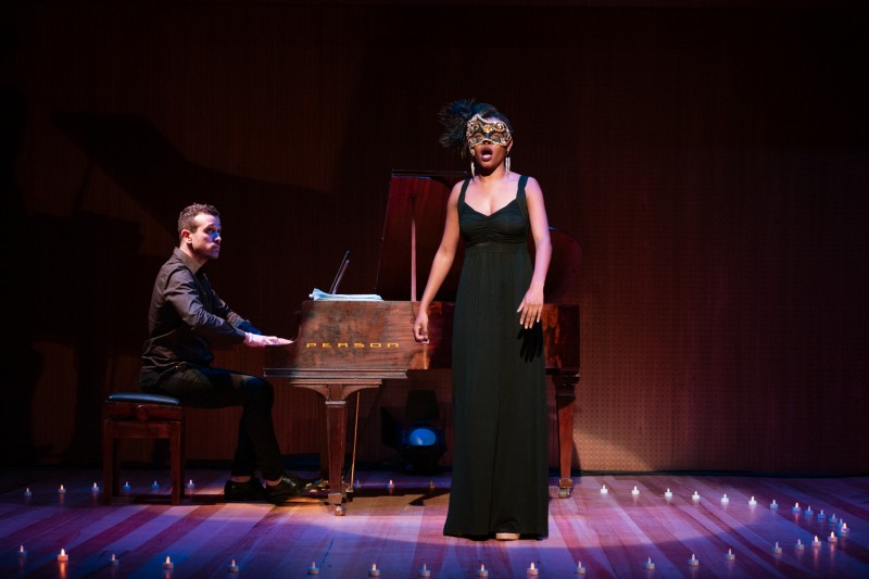 No palco de um teatro, uma mulher negra de vestido preto está de pé e traja uma máscara que cobre os olhos. Ela canta. Ao fundo, um homem toca piano. O cenário é de cor mais escura.