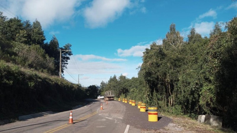 Imagem do trecho de estrada que foi liberado. A imagem mostra um trecho em curva com árvores dos dois lados e tambores amarelos de sinalização do lado direito.