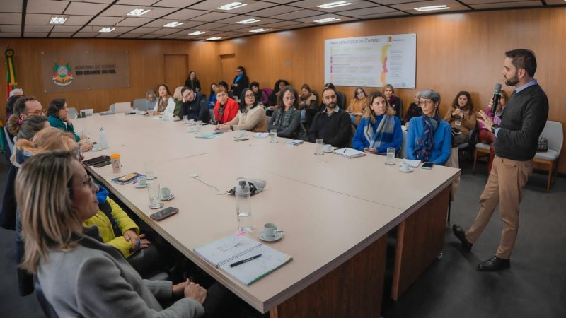 O vice-governador Gabriel Souza está em pé, à direita da imagem, falando ao microfone para os participantes do encontro, que se encontram sentados diante de uma mesa retangular grande. Há ainda pessoas sentadas em um fileira de cadeiras que pode ser vista atrás das pessoas que estão à mesa.