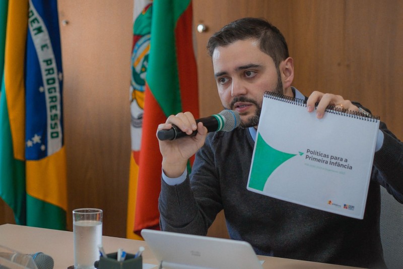 Gabriel Souza sentado falando ao microfone enquanto segura na mão esquerda um documento intitulado Políticas para a Primeira Infância