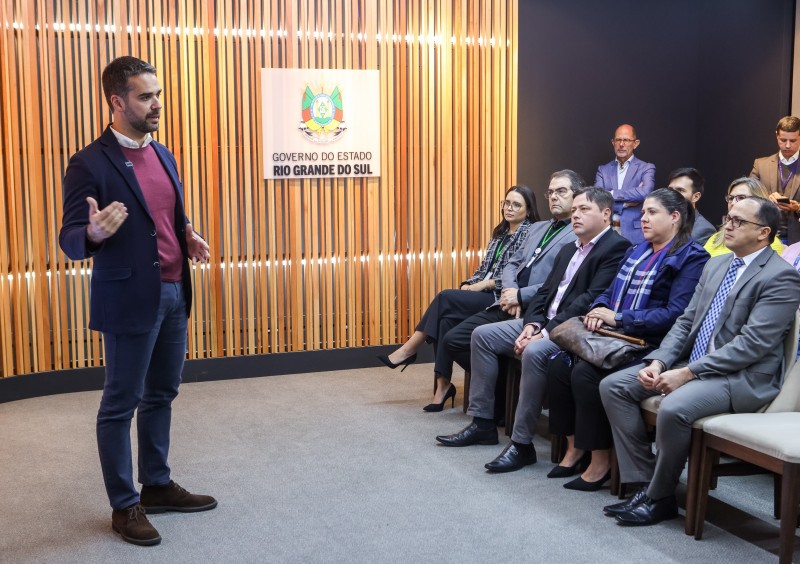 Leite em pé mais à esquerda da imagem falando para uma plateia. No centro da imagem, há uma placa com o brasão do Rio Grande do Sul pregada na parede. Na parte direita inferior da imagem aparecem pessoas sentadas na plateia.