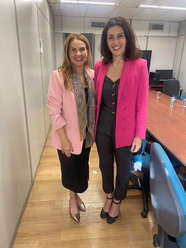 Numa sala de reuniões, Leany e Simone posam para a foto, de pé. Ambas sorriem. A sala tem uma grande mesa e algumas cadeiras de escritório.