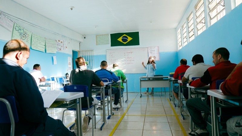 Sala de aula vista pelos fundos. Uma professora dá aula para os estudantes, sentados em suas classes. Todos os alunos são homens adultos