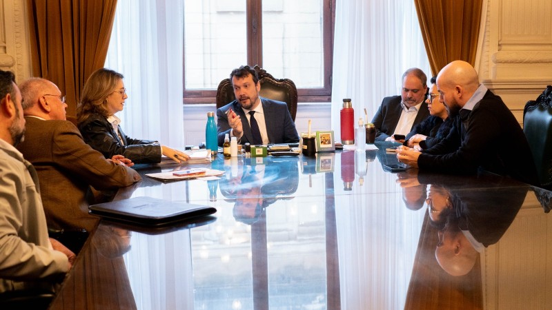 Numa sala de reuniões, secretários estão sentados à mesa, que é grande e de tampo com reflexo. São 7 pessoas, uma delas está na cabeceira e aparece de frente. Atrás, uma grande janela