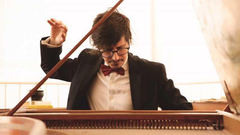 Fernando Cordella de frente tocando cravo e regendo uma orquestra ao mesmo tempo.