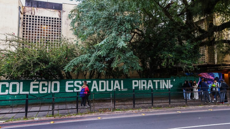 Fachada do Colégio Estadual Piratini. Na frente dele, um grupo de alunos conversa e um pouco mais distante dois alunos se abraçam.