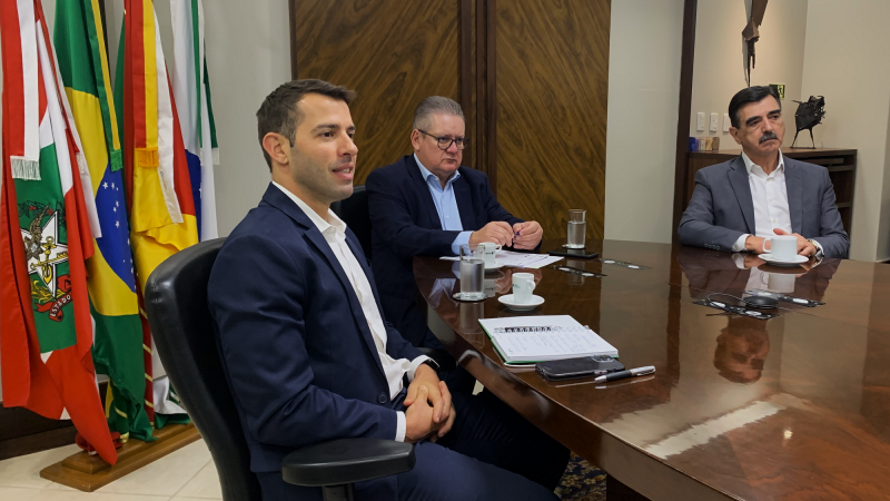 Busatto participando da primeira reunião da diretoria do BRDE sentado à mesa acompanhado de mais duas pessoas. Todos olham para uma tela enquanto dialogam com outros diretores que participam de modo virtual da reunião.