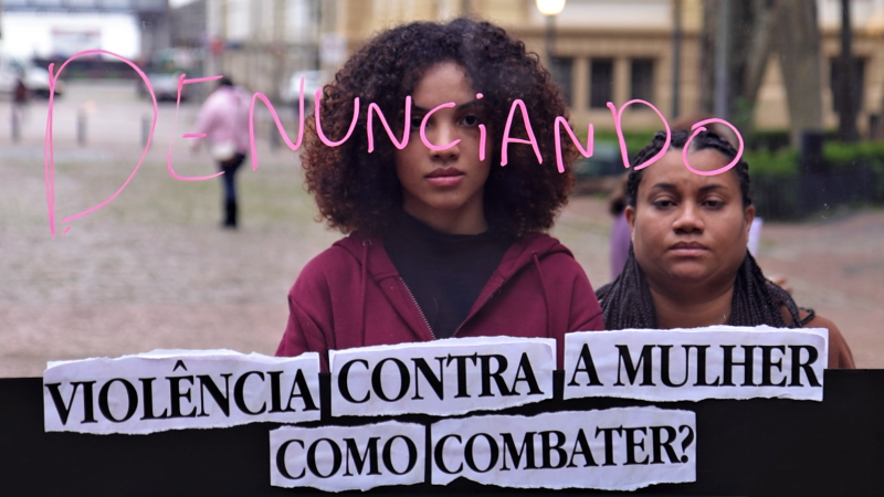 Duas mulheres negras atrás do painel transparente no qual está escrita a palavra denunciando como a resposta ao que está escrito na base do painel: violência contra mulher, como combater? Ao fundo se vê a Praça da Alfândega desfocada na imagem.