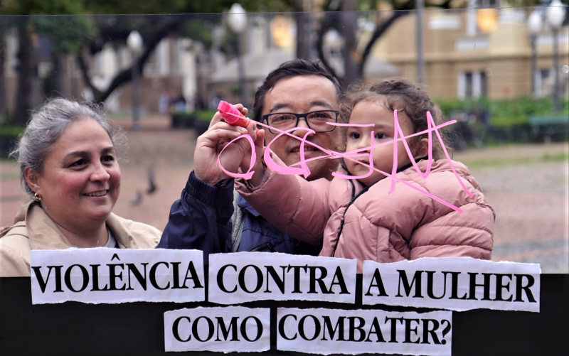 Uma mulher e um homem com uma menina no colo atrás do painel transparente no qual está escrita a palavra medo com dois riscos cruzados sobre ela como a resposta ao que está escrito na base do painel: violência contra mulher, como combater? Ao fundo se vê a Praça da Alfândega desfocada na imagem.