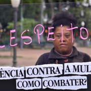 Uma mulher atrás do painel transparente no qual está escrita a palavra respeito como resposta ao que está escrito na base do painel: violência contra mulher, como combater? Ao fundo se vê a Praça da Alfândega desfocada na imagem.