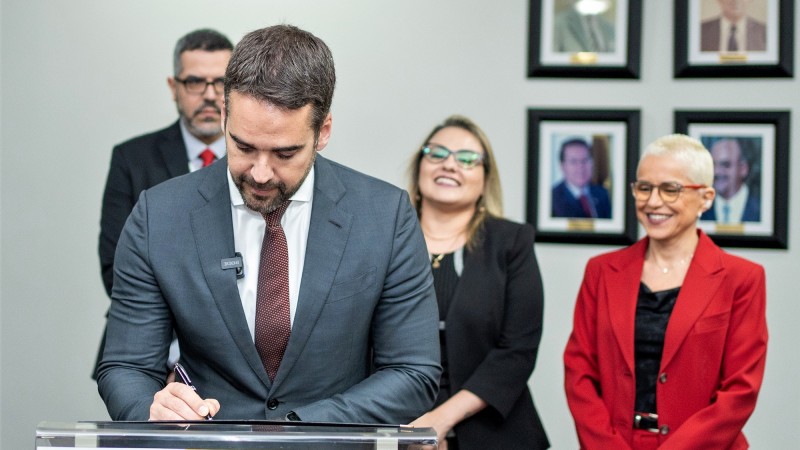Imagem do governador Eduardo Leite assinando o termo que se encontra sobre um púlpito com a logomarca dos Correios. Ao fundo podem ser vistas três pessoas envolvidas na assinatura. 