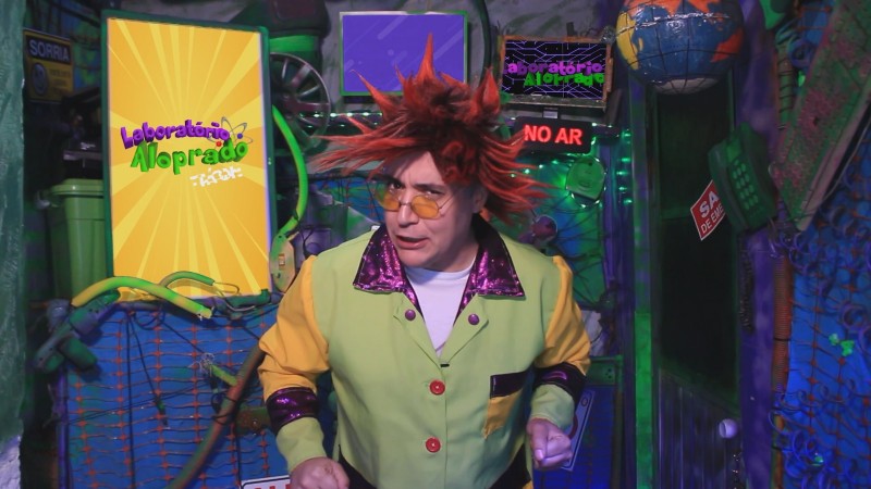 Um ator do programa aparece caracterizado como um cientista, trajando um jaleco de cores exageradas (amarelo, verde e roxo). Seu cabelo é vermelho e espetado, e ele usa óculos de lentes laranjas. Ele encara a câmera com olhar arguto. No fundo, aparece seu laboratório, bastante bagunçado e colorido. À esquerda, há uma tela com a logo do programa.