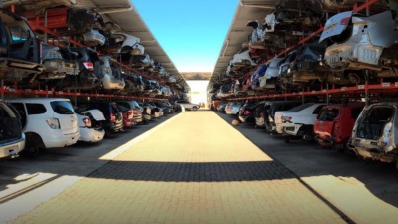 Foto de um desmanche regularizado. Há uma rua no centro e dois galpões abertos à esquerda e à direita com sucatas de carros dispostos em três andares.