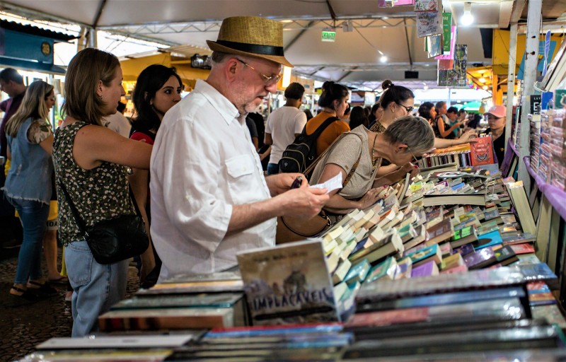 Imagem de uma banca livros com pessoas buscando volumes. Ao fundo se vê imagens da feira do livro, com outras bancas e pessoas andando pelo corredor.