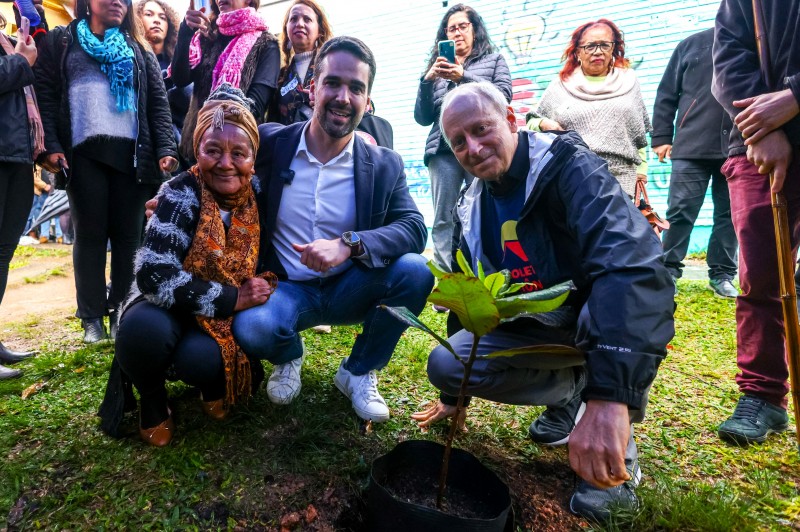 Leite ao centro da imagem abraçado com uma mulher negra. À direita está Sandel. Os três estão agachados plantando uma muda de árvore no terreno da escola. Ao fundo, pessoas obaservam a cena.