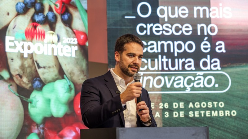 Em ambiente fechado, governador Eduardo Leite fala no microfone, no lançamento da Expointer 2023. Ao fundo, imagens da campanha da feira deste ano, com destaque para a frase "O que mais cresce no campo é a cultura da inovação".