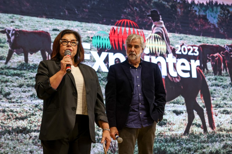 Em frente ao painel com a marca da Expointer 2023, em ambiente fechado, aparecem Tânia e Beto. A secretária Tânia fala ao microfone no lançamento da Expointer 2023.