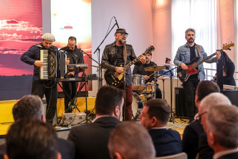 Em frente ao painel com a marca da Expointer 2023, músicos tocando teclado, gaita, guitarra e bateria se apresentam no lançamento da feira.