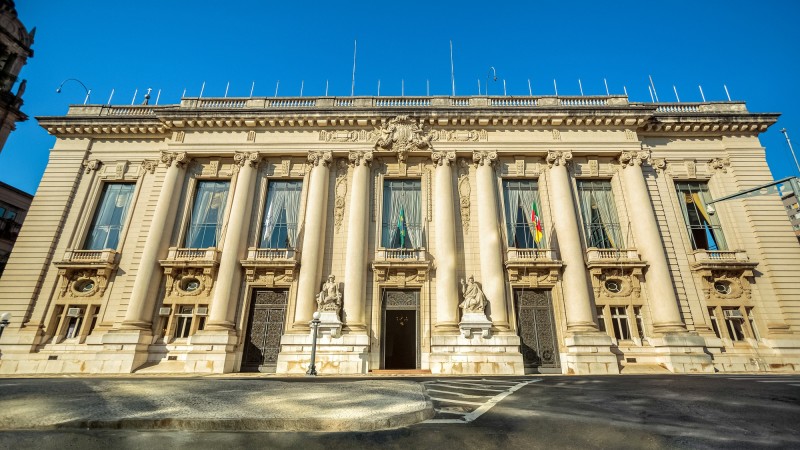 Vista frontal do Palácio Piratini. A parte da frente do palácio possui grandes janelas, uma portal de acesso ao meio, as bandeiras de Brasil e do RS, algumas estátuas e grandes pilares. O céu está azul.