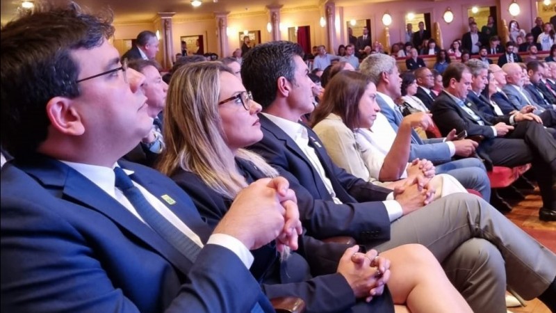 Várias pessoas sentadas em na plateia de um grande teatro, atentas ao que está se passando no palco.  