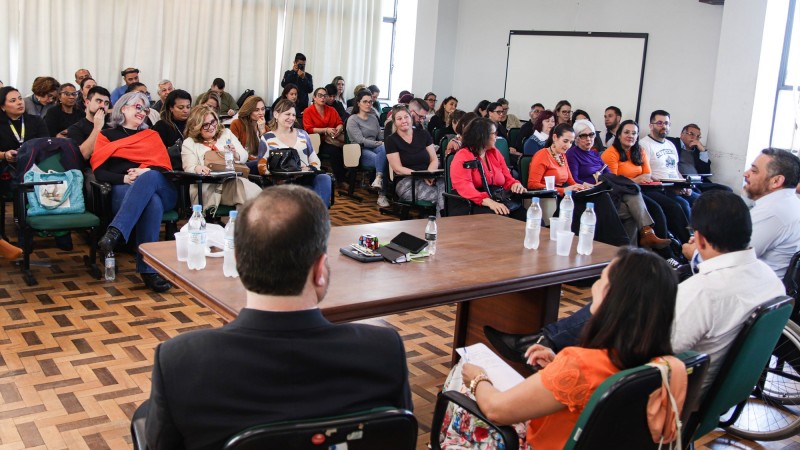 Na imagem pode ser vista uma plateia de frente assistindo às falas dos componentes da mesa, que são vistos de costas mais à direita.