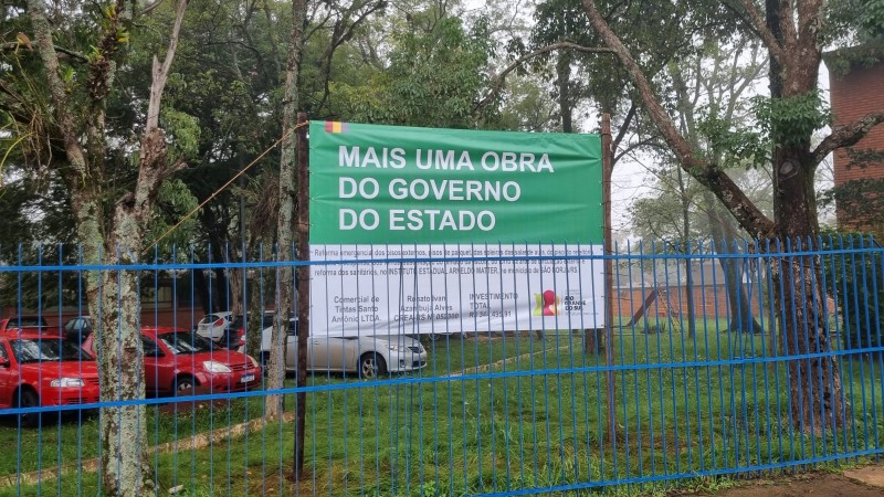 Fincada no gramado da entrada da escola, há uma grande placa onde está escrito "Mais uma obra do governo do Estado", com letras brancas em fundo verde. Na metade inferior da placa, de cor branca, há escrito pequenos e a logo do governo estadual. Na foto, é possível ver ainda árvores e o estacionamento da escola.