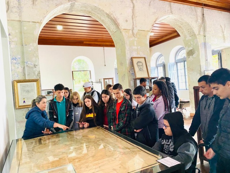 No museu, que possui pilastras brancas de pedra e teto de madeira, um grupo de estudantes em idade escolar acompanha a fala de uma guia, que aponta um mostruário de vidro onde está um mapa antigo.