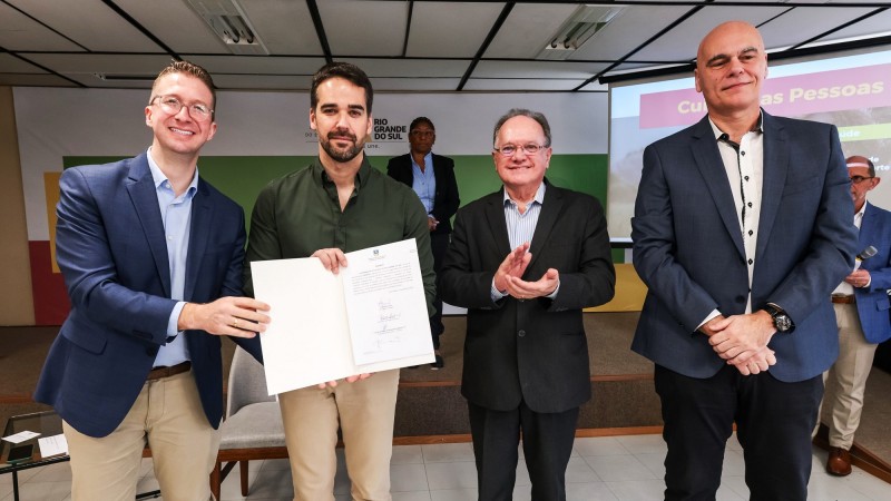 Eduardo Leite ao centro e Fantinel mais à esquerda da imagem segurando o documento assinado que cria o programa Universitário do Amanhã. Há mais dois homens na foto. Um ao centro e ao lado de Leite e outro mais à direita da imagem.