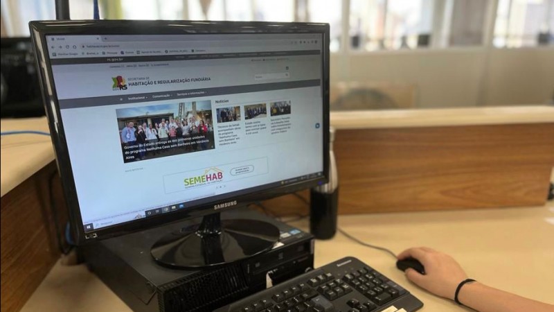Em close, mais à esquerda da imagem, há uma tela de computador sobre uma cpu e o teclado. Os braços de uma pessoas estão sobre a mesa e uma delas manuseia um mouse mais à direita da imagem.