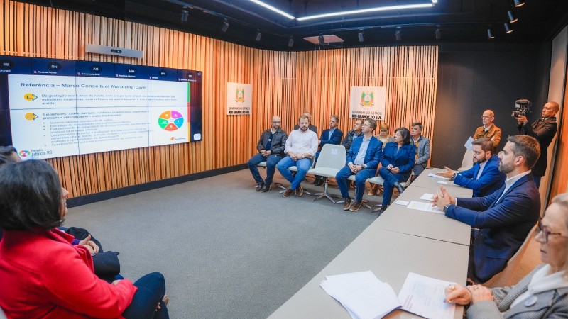 Numa sala fechada, várias pessoas estão sentadas no formato de U, assistindo a um telão com a apresentação, que aparece à esquerda. Na direita, Leite e Gabriel realizam a apresentação.
