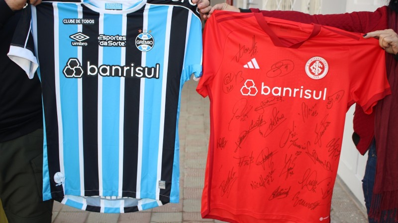 Uma de cada lado, duas pessoas exibem as camisetas oficiais de Grêmio e Inter. A do primeiro, é azul, preto e branco; a do segundo é vermelha.