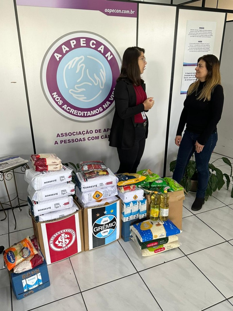 Em frente a um painel com o símbolo da AAPECAN, que é um círculo roxo e azul claro com três mãos juntas no meio, estão os alimentos empilhados. Em duas caixas, estão o símbolo do Internacional e do Grêmio. Há duas mulheres em pé ao lado dos alimentos. Elas conversam entre si.