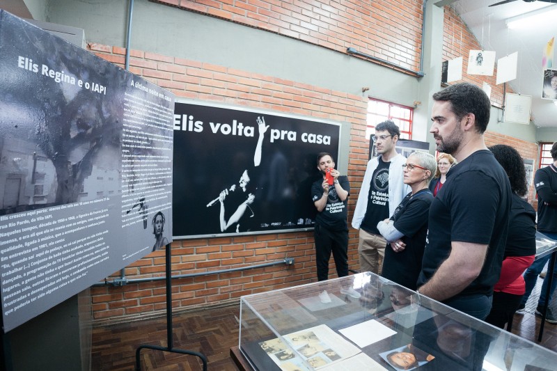 O governador Eduardo Leite e a secretária da Cultura, Beatriz Araújo, ambos vestindo camisetas pretas, observam painéis com fotos e textos da exposição Elis Volta para Casa na Biblioteca Romano Rief.