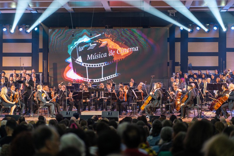 Músico da Ospa no palco da Casa da Música. Ao fundo, um letão com uma arte que leva a ilustração de um microfilme e o texto Música de Cinema. Em primeiro plano na imagem, a plateia que lotou o espaço para assistir o espetáculo.
